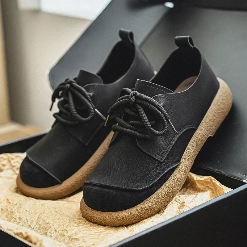Paire de chaussures noires à semelle crêpe beige, présentée dans une boîte noire sur papier froissé. 12