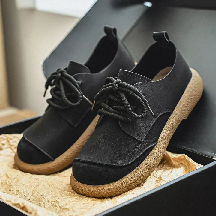 Paire de chaussures noires à semelle crêpe beige, présentée dans une boîte noire sur papier froissé. 12