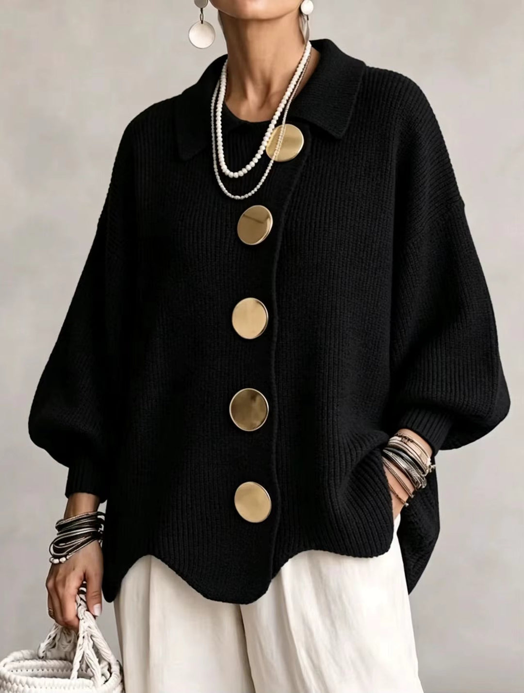 Vue de face du cardigan noir en maille côtelée, grands boutons dorés asymétriques, bas ondulé, porté avec un pantalon blanc. 3