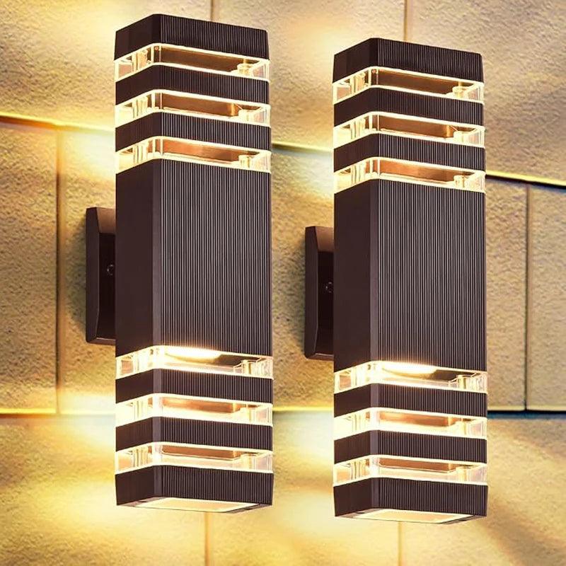 Deux appliques rectangulaires noires à rainures verticales et bandes transparentes, allumées en lumière chaude, fixées sur un mur de pierre. 1