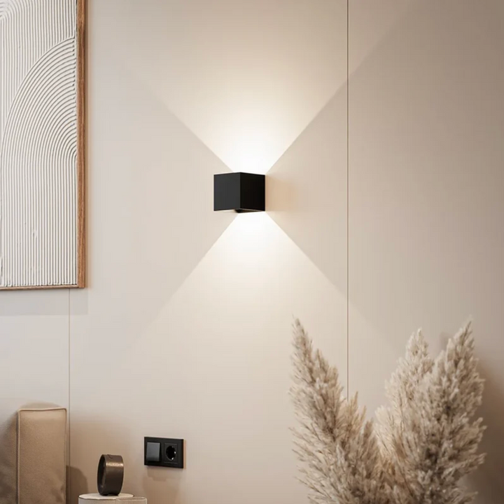 Applique cubique noire allumée sur un mur beige, projetant un faisceau de lumière vers le haut et le bas, près d'herbes de la pampa. 1