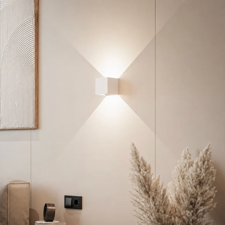 Applique cubique blanche allumée sur un mur beige, avec cadre décoratif, herbes de la pampa et prise murale noire à proximité. 12