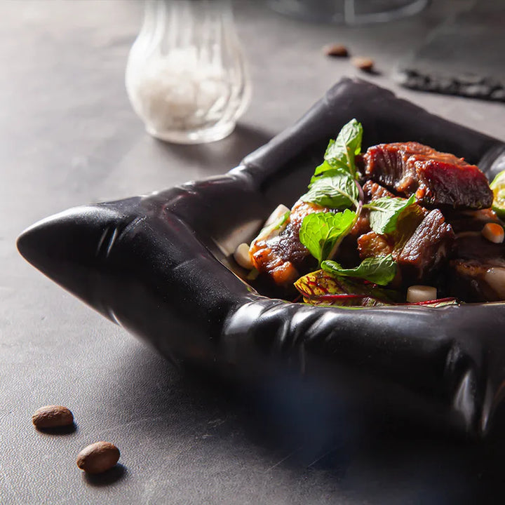 Gros plan sur un angle du saladier noir brillant aux plis marqués, garni de viande et de feuilles de menthe, avec une salière floue. 4