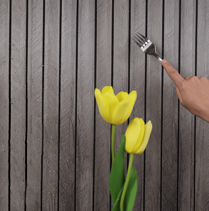 Mur de lattes grises texturées, une main pointant une fourchette vers la surface, avec deux tulipes jaunes au premier plan. 7