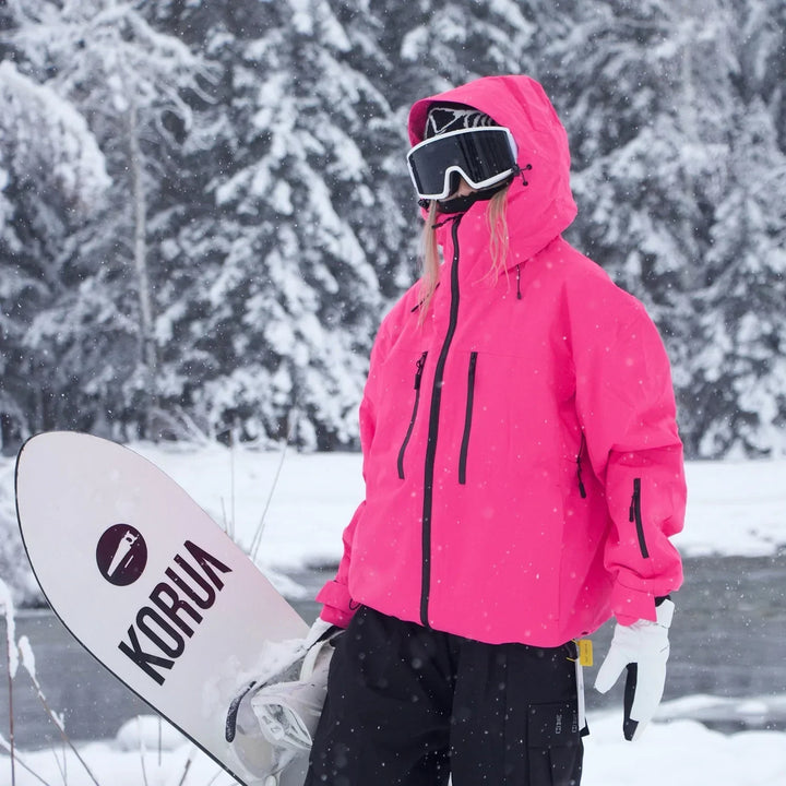 Veste rose fuchsia vif à capuche portée par une personne tenant un snowboard blanc, avec pantalon noir et gants blancs dans la neige. 5