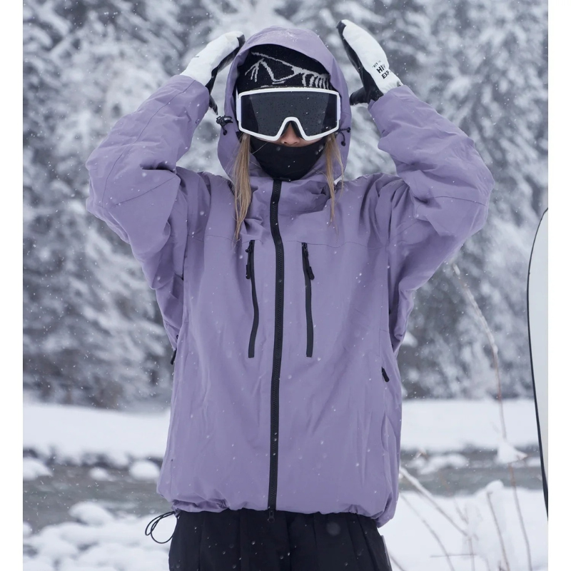 Veste violet lavande portée par une personne qui remonte la capuche des deux mains, masque de ski sur le visage, forêt enneigée en fond. 1