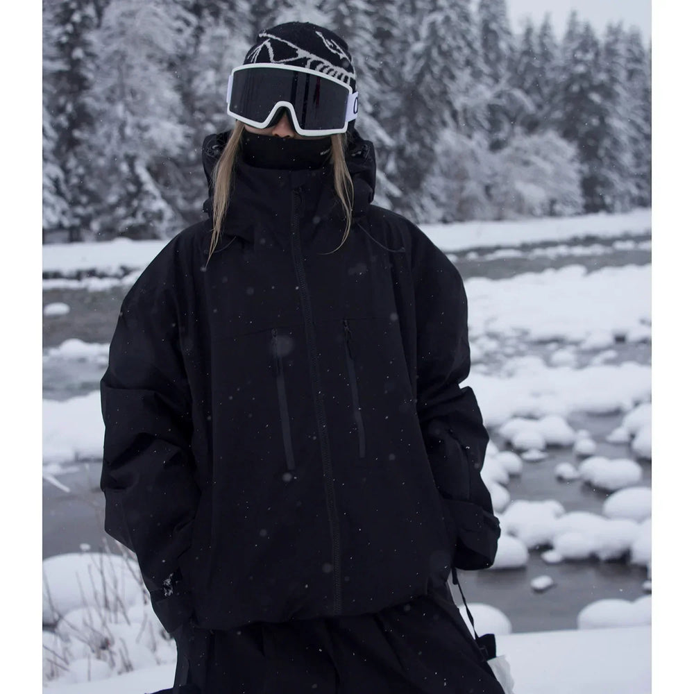 Veste noire à capuche portée en pied par une personne avec bonnet, masque de ski et tour de cou, au bord d'une rivière enneigée. 2