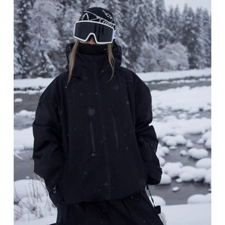 Veste noire à capuche portée en pied par une personne avec bonnet, masque de ski et tour de cou, au bord d'une rivière enneigée. 2