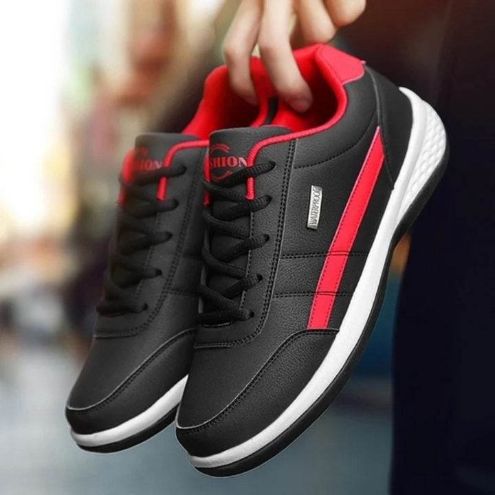 Paire de baskets noires à bande latérale rouge et semelle blanche, tenue à la main devant un décor de rue flou. 1