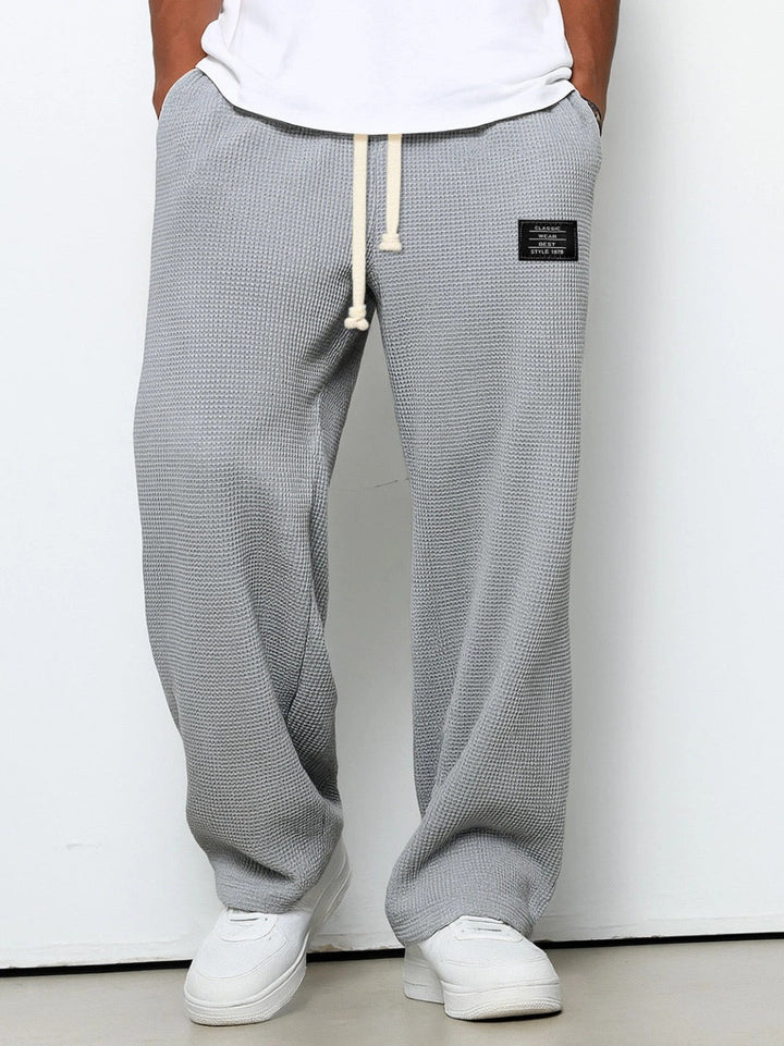 Version gris moyen du pantalon large en maille gaufrée, cordon de serrage beige, portée par un modèle devant un mur clair. 3