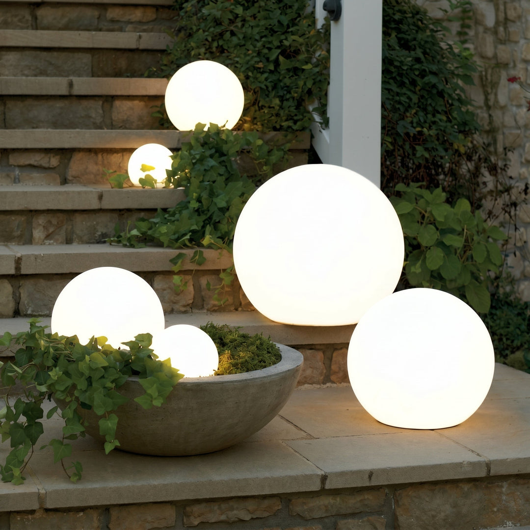 Six sphères lumineuses blanches de tailles variées posées sur des marches en pierre et dans une vasque, entourées de lierre, à la tombée de la nuit. 1