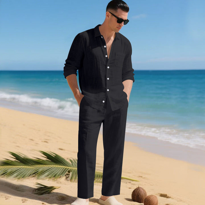 Homme sur une plage portant l'ensemble chemise à manches longues et pantalon droit en noir, mains dans les poches. 1