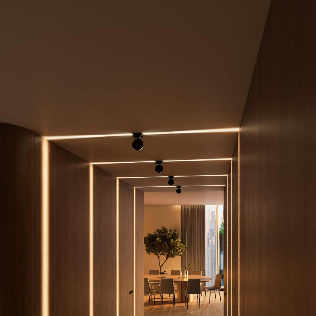 Couloir en bois avec quatre appliques noires alignées au plafond, faisceaux horizontaux chauds et salle à manger en arrière-plan. 14