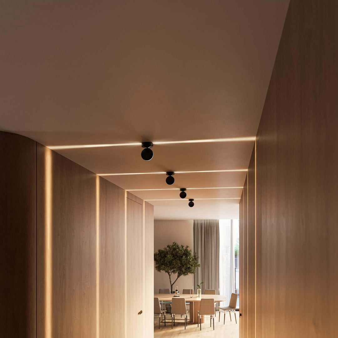 Appliques noires alignées au plafond d'un couloir en bois, faisceaux lumineux doux et table à manger visible au fond. 17