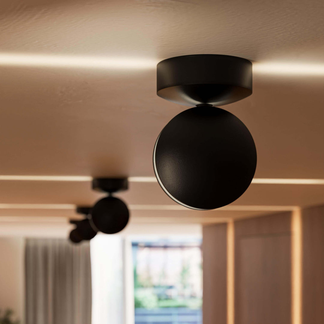 Gros plan sur une applique noire mate au plafond, tête ronde pivotante, avec des lignes de lumière chaude sur le plafond. 11