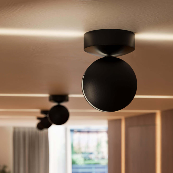 Gros plan sur une applique noire mate au plafond, tête ronde pivotante, avec des lignes de lumière chaude sur le plafond. 11