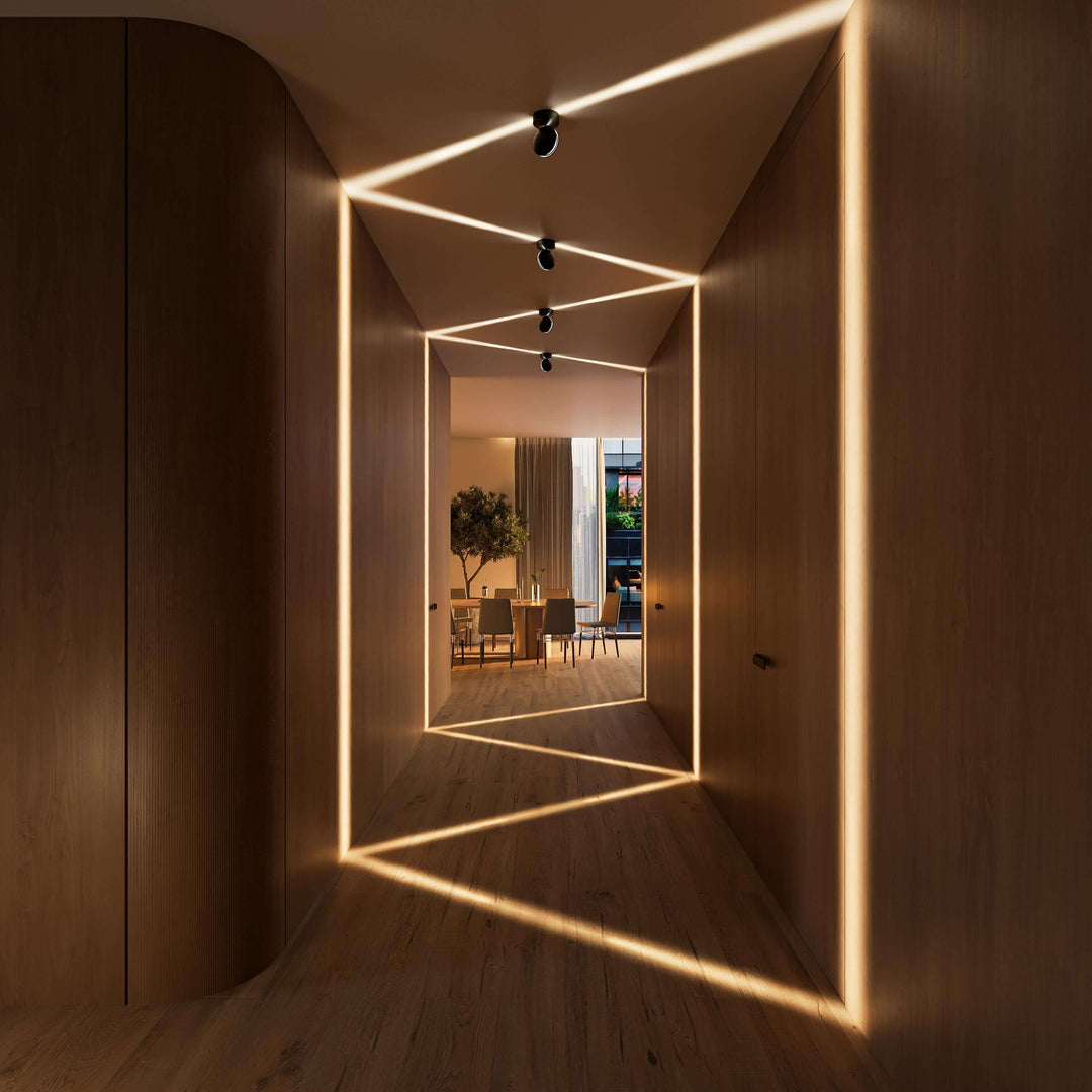 Couloir en bois avec appliques noires au plafond projetant des faisceaux en zigzag sur les murs et le parquet, salle à manger au fond. 10