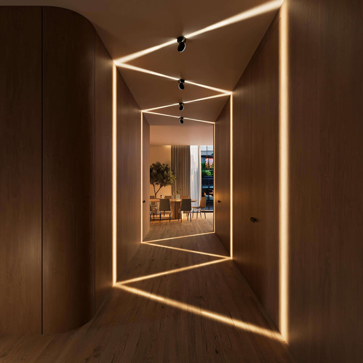 Couloir en bois avec appliques noires au plafond projetant des faisceaux en zigzag sur les murs et le parquet, salle à manger au fond. 10