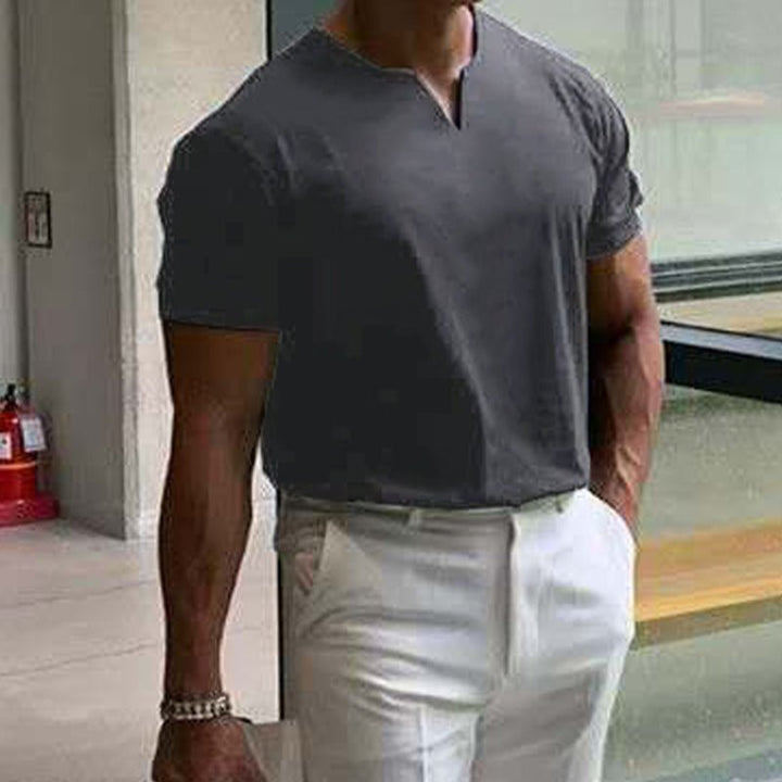 T-shirt gris foncé à col V porté avec un pantalon blanc, main dans la poche, dans un couloir lumineux. 5