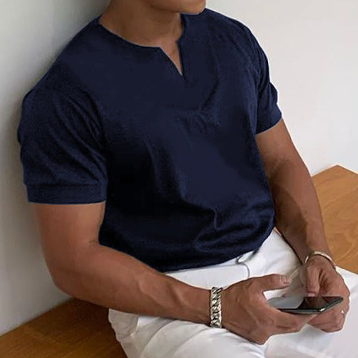 T-shirt bleu marine à col V porté par un homme assis sur un banc en bois, pantalon blanc, smartphone en main. 6