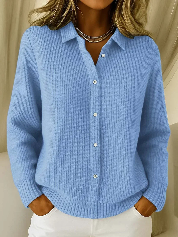 Femme portant le pull en maille texturée bleu ciel, col chemise et boutons blancs, mains dans les poches d'un pantalon blanc. 1