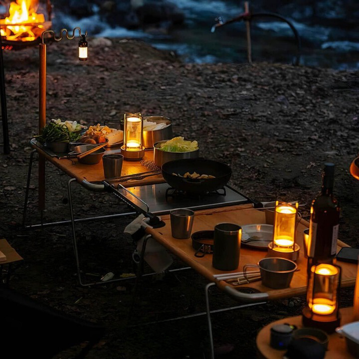 Plusieurs lampes allumées sur des tables de camping en bois, avec casseroles et repas, au bord d'une rivière le soir. 4
