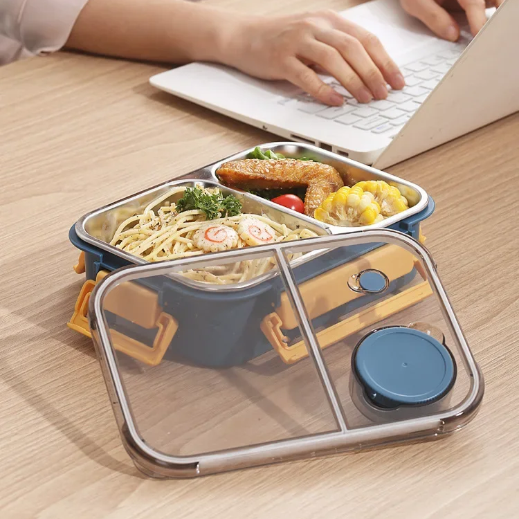 Boîte bleu et jaune garnie de spaghettis, poulet et maïs, posée sur un bureau devant un ordinateur portable, avec son couvercle transparent. 4