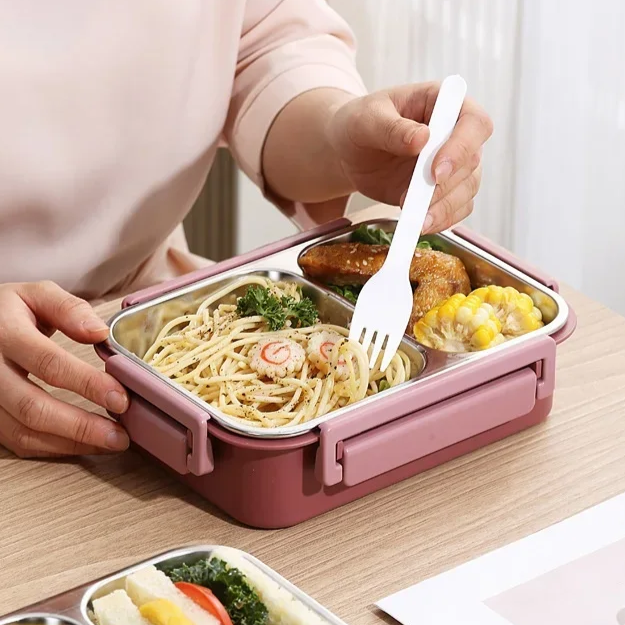 Une personne mange des spaghettis avec une fourchette blanche dans une boîte repas rose à compartiments, posée sur une table en bois. 5