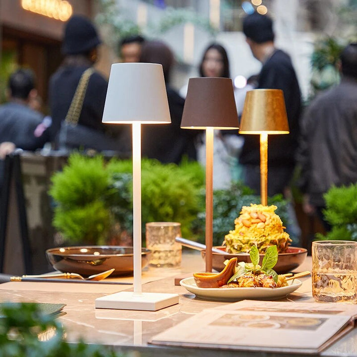 Trois lampes allumées, blanche, brune et dorée, posées sur une table de restaurant parmi des assiettes et des verres. 2