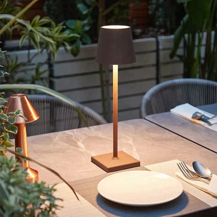 Lampe cuivrée allumée sur une table de terrasse en marbre, entourée de plantes, avec une assiette et des couverts au premier plan. 3