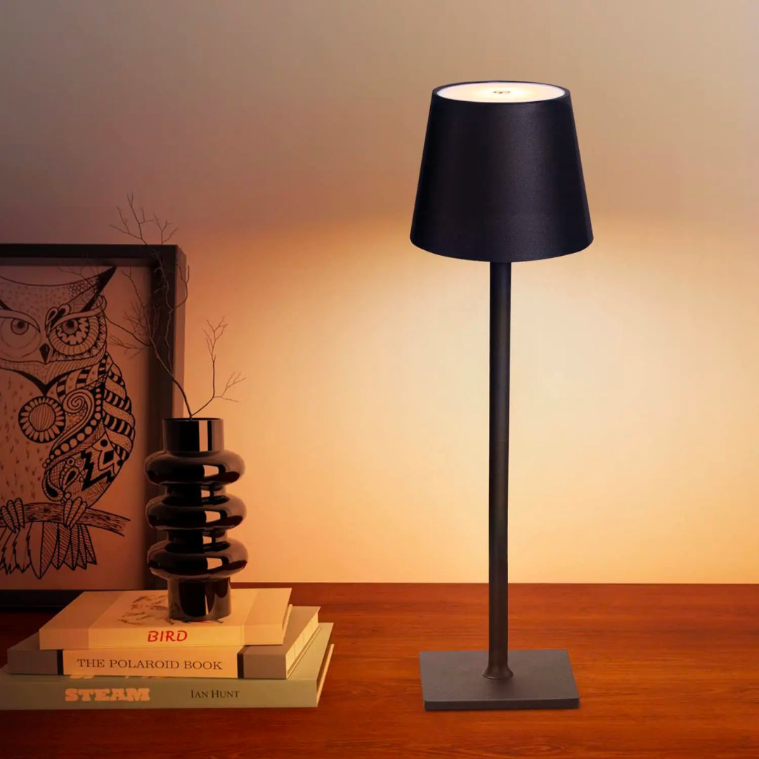 Lampe noire allumée sur une table en bois, près d'une pile de livres, d'un vase en céramique et d'un cadre avec un hibou. 1