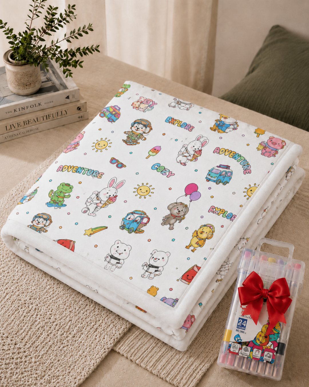 Plaid plié blanc orné de petits animaux aventuriers déjà colorés, avec un coffret de marqueurs et ruban rouge 10