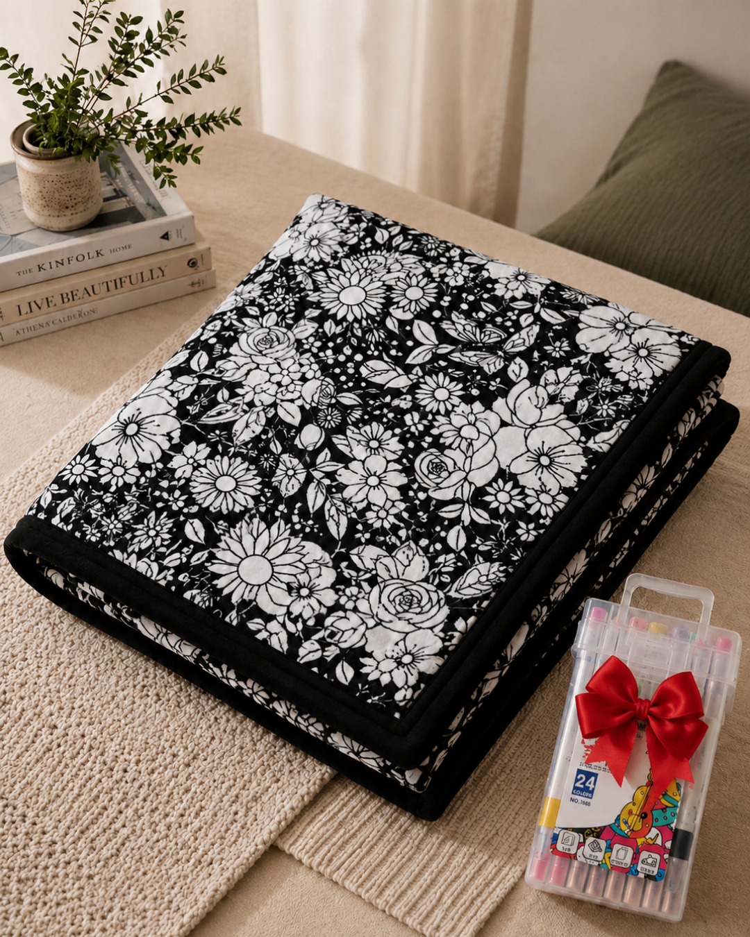 Couverture pliée à motif floral blanc sur fond noir, posée avec un coffret de feutres noué d'un ruban rouge 1