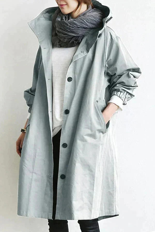 Trench gris clair à capuche porté de face, boutonné, mains dans les poches, avec écharpe grise et haut blanc. 3