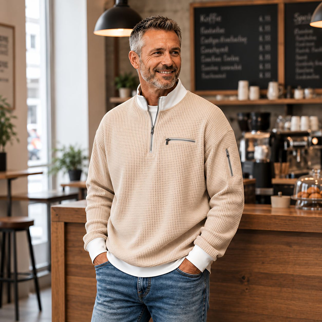 Homme souriant en pull beige gaufré à col zippé, mains dans les poches de son jean, dans un café. 1