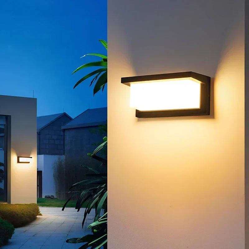 Applique noire à lumière chaude fixée sur un mur clair, devant un jardin avec plantes et maisons 4