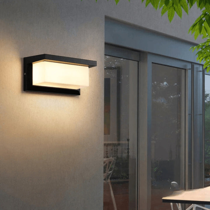 Applique rectangulaire à cadre noir allumée en lumière chaude sur un mur crépi, près de baies vitrées 1