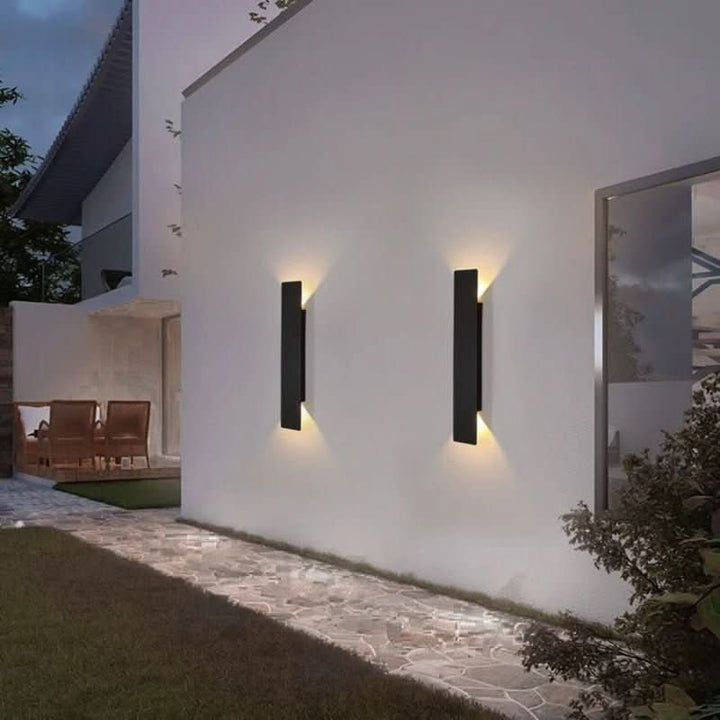 Deux appliques noires allongées sur un mur blanc extérieur le soir, avec un chemin en pierre et un coin salon en arrière-plan. 3