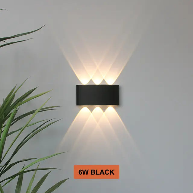Applique noire à trois faisceaux haut et trois bas, allumée sur un mur gris, plante verte à gauche. 11
