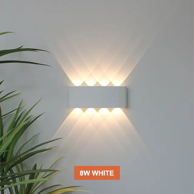 Applique blanche à quatre faisceaux haut et quatre bas, lumière chaude croisée sur un mur clair, plante verte à gauche. 12