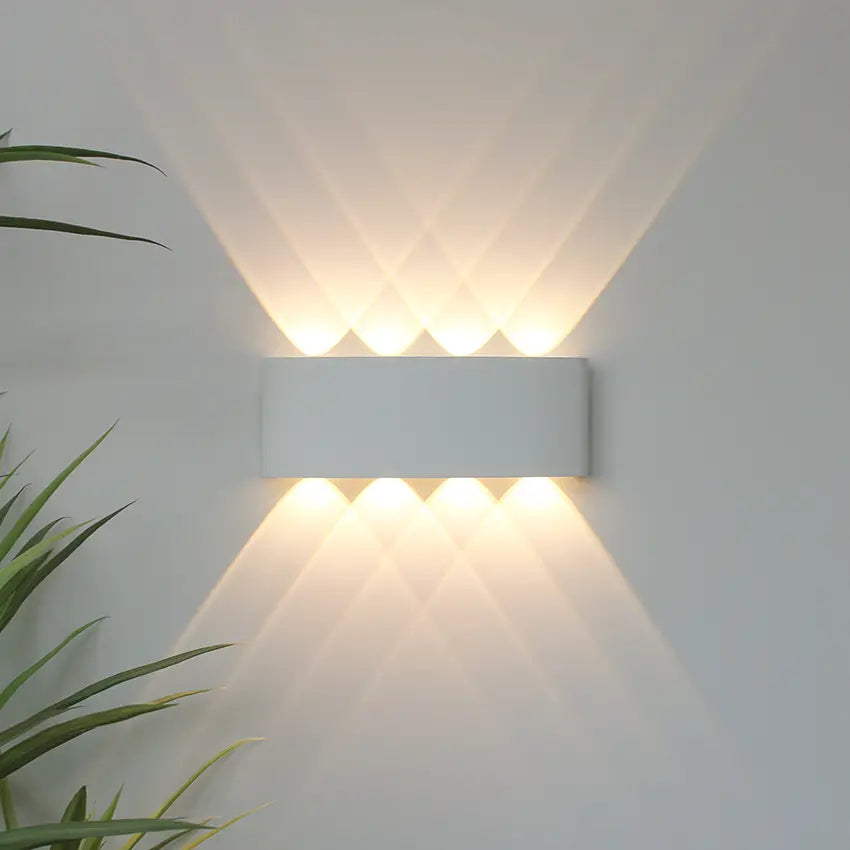 Applique blanche allumée sur un mur clair, projetant huit faisceaux de lumière chaude croisés vers le haut et le bas, plante verte à gauche. 1
