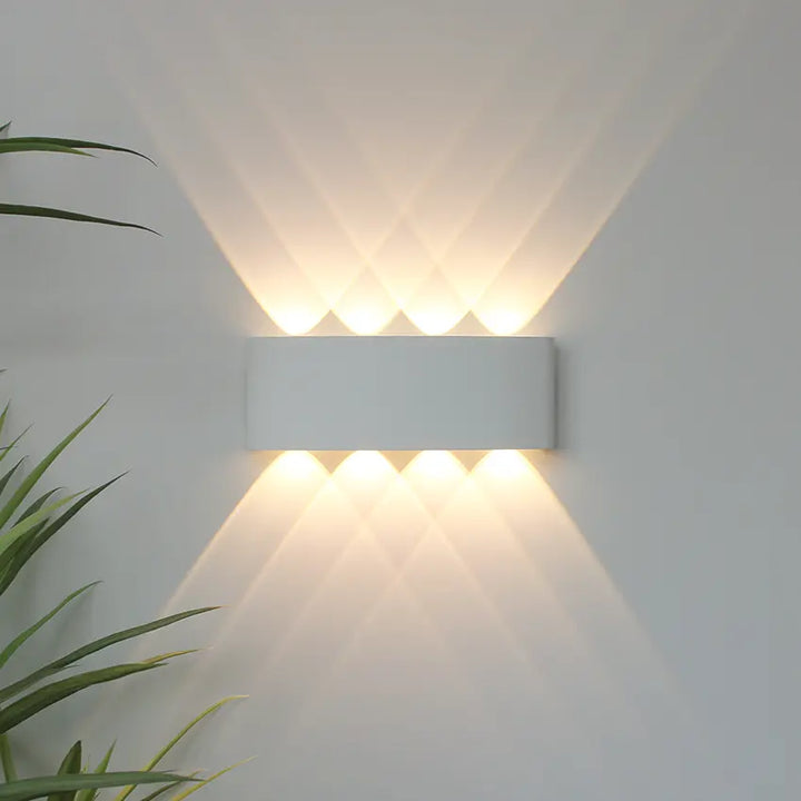 Applique blanche allumée sur un mur clair, projetant huit faisceaux de lumière chaude croisés vers le haut et le bas, plante verte à gauche. 1