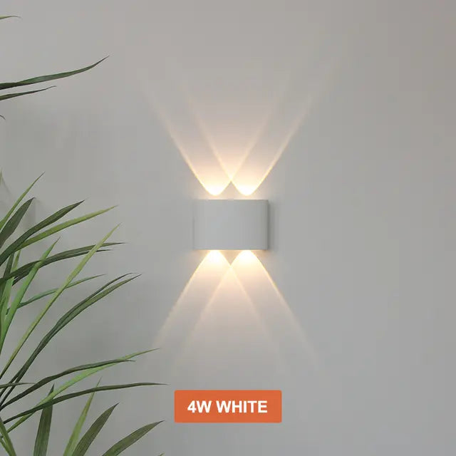 Applique blanche à deux faisceaux haut et deux bas, allumée sur un mur clair à côté d'une plante verte. 6