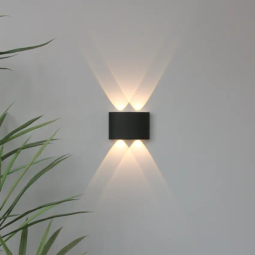 Applique noire à deux faisceaux haut et deux bas, lumière chaude croisée sur un mur gris, feuilles vertes à gauche. 7