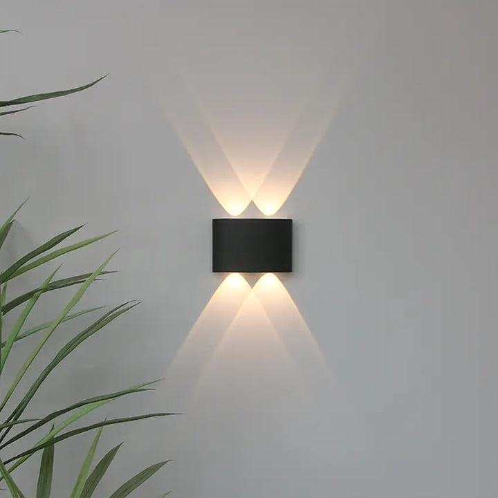 Applique noire à deux faisceaux haut et deux bas, lumière chaude croisée sur un mur gris, feuilles vertes à gauche. 7