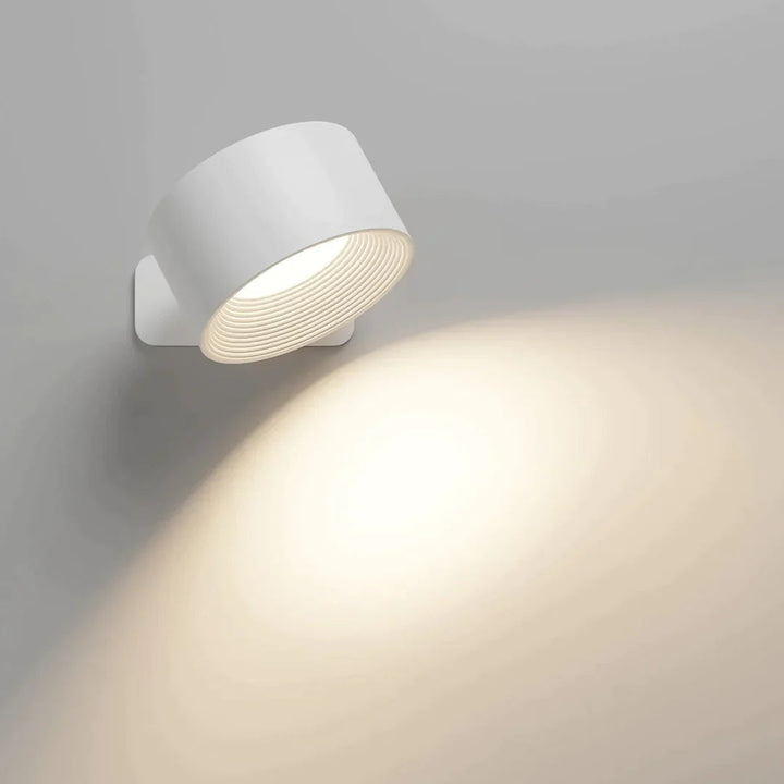 Applique cylindrique blanche allumée, inclinée vers le bas, projetant un cône de lumière chaude sur un mur gris clair. 1
