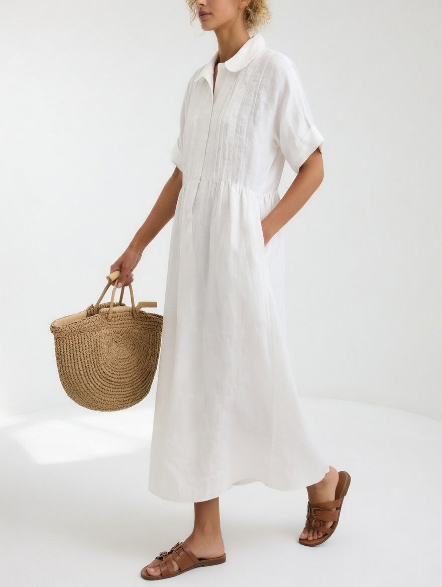 Femme de profil portant la robe blanche à col rond et plis sur le buste, une main dans la poche, panier en paille. 1