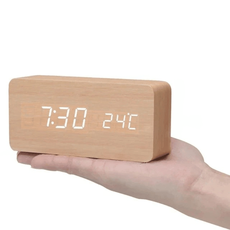 Réveil rectangulaire en bois clair posé sur une main ouverte, affichage LED blanc de l'heure et de la température. 15
