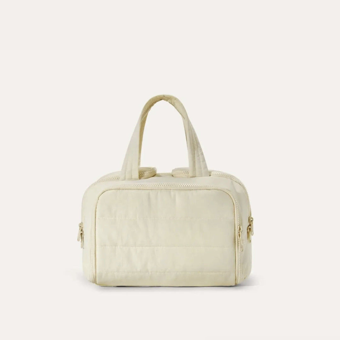 Trousse matelassée de face dans un coloris crème, avec anses souples et fermetures éclair dorées sur les côtés. 16