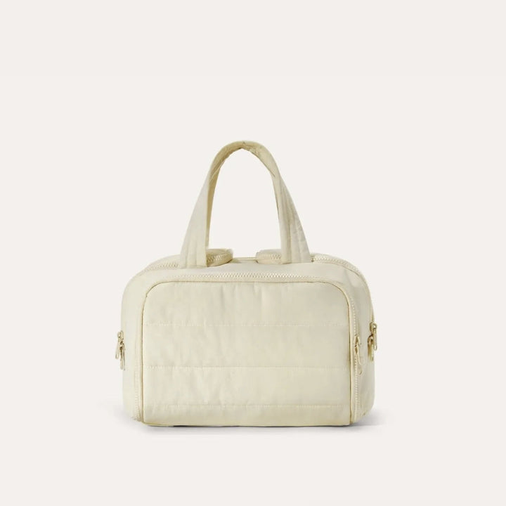 Trousse matelassée de face dans un coloris crème, avec anses souples et fermetures éclair dorées sur les côtés. 16